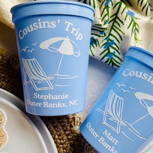 May include: Two light blue plastic cups with white text and graphics. The cups read "Cousins' Trip" with a beach umbrella, a lounge chair, and the name "Stephanie Outer Banks, NC" on one cup and "Matt Outer Banks" on the other.