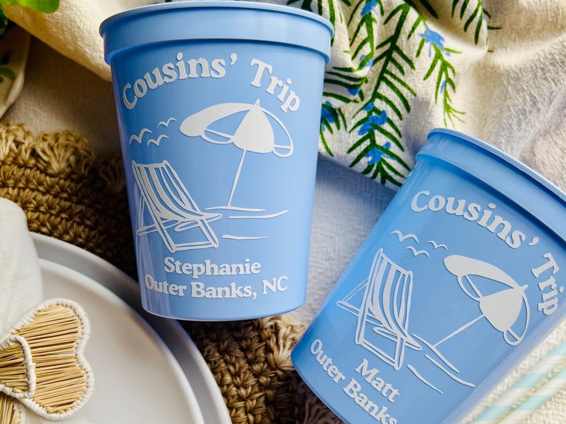 May include: Two light blue plastic cups with white text and graphics. The cups read "Cousins' Trip" with a beach umbrella, a lounge chair, and the name "Stephanie Outer Banks, NC" on one cup and "Matt Outer Banks" on the other.