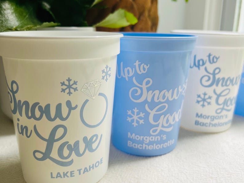 May include: White and light blue plastic cups with text and snowflake designs. The cups have phrases like "Snow in Love" and "Up to Snow Good". The text "Lake Tahoe" and "Morgan's Bachelorette" are also visible.