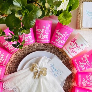 May include: A festive table setting featuring pink cups and napkins with the text "Snow in Love" and a snowflake design. The cups are arranged around a white plate with a gold bow napkin ring. Green foliage and white flowers add a natural touch.