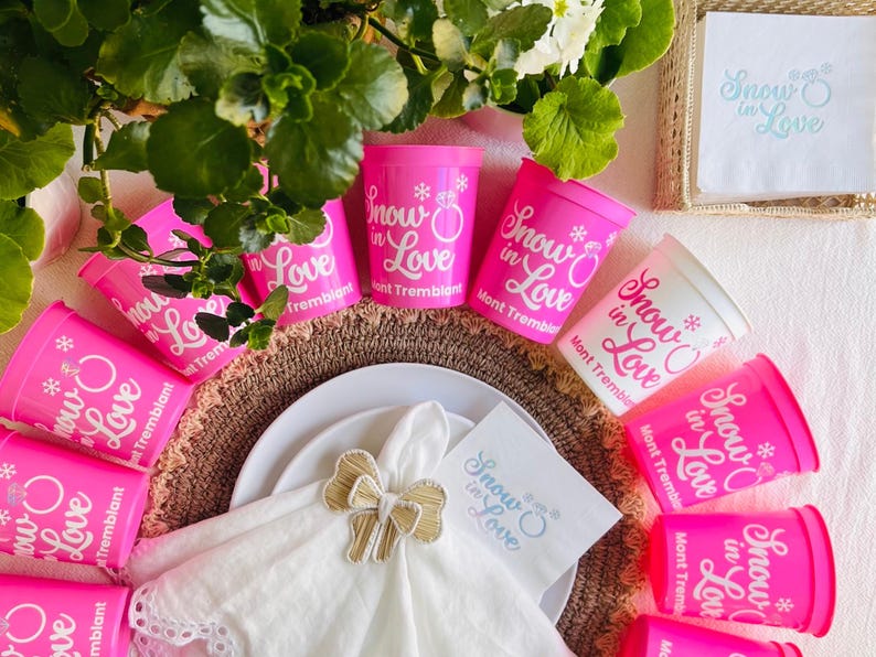 May include: A festive table setting featuring pink cups and napkins with the text "Snow in Love" and a snowflake design. The cups are arranged around a white plate with a gold bow napkin ring. Green foliage and white flowers add a natural touch.