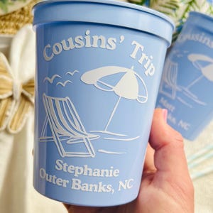 Hand holds a light blue stadium cup that is made for a beach trip. The cup features a graphic of a beach chair beside an umbrella with waves and seagulls in the background of the image. Two lines of customizable text appear below the design, and one line of custom text arcs over the design.