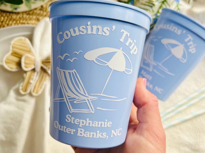 Hand holds a light blue stadium cup that is made for a beach trip. The cup features a graphic of a beach chair beside an umbrella with waves and seagulls in the background of the image. Two lines of customizable text appear below the design, and one line of custom text arcs over the design.