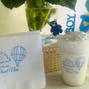 Op de afbeelding: Witte papieren servetten met een blauw ontwerp van een luchtballon en een wolk. De tekst "We are on Cloud Nine" staat op de servet gedrukt. Een blauwe teddybeer, een blauw "Boy"-bord en een blauwe romper zijn op een witte, geglazuurde cupcake.