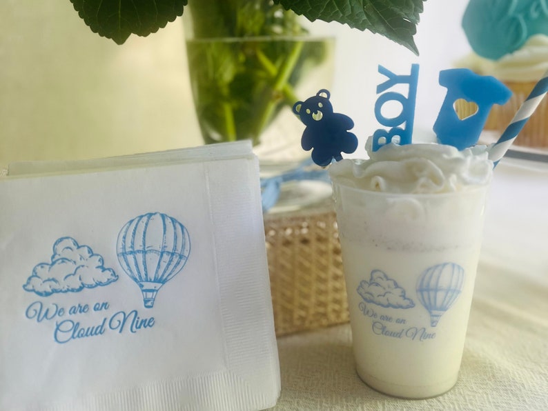 Op de afbeelding: Witte papieren servetten met een blauw ontwerp van een luchtballon en een wolk. De tekst "We are on Cloud Nine" staat op de servet gedrukt. Een blauwe teddybeer, een blauw "Boy"-bord en een blauwe romper zijn op een witte, geglazuurde cupcake.