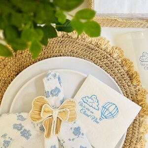 Op de afbeelding: Een witte servet met blauwe bloemenprint is vastgebonden met een natuurlijke geweven strik. Een witte servet met blauwe tekst die "We are on Cloud Nine" zegt en een blauwe illustratie van een wolk en een luchtballon ligt op een beige placemat.