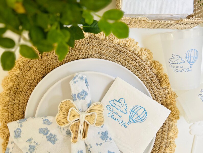 Op de afbeelding: Een witte servet met blauwe bloemenprint is vastgebonden met een natuurlijke geweven strik. Een witte servet met blauwe tekst die "We are on Cloud Nine" zegt en een blauwe illustratie van een wolk en een luchtballon ligt op een beige placemat.