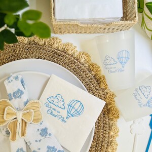 Op de afbeelding: Een witte servet met blauwe tekst die "We are on Cloud Nine" zegt en een blauwe illustratie van een luchtballon en een wolk. Een bruine geweven servetring is vastgebonden aan een witte servet met een blauw bloemenpatroon.