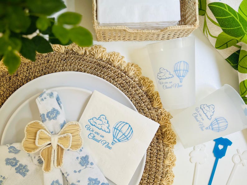 Op de afbeelding: Een witte servet met blauwe tekst die "We are on Cloud Nine" zegt en een blauwe illustratie van een luchtballon en een wolk. Een bruine geweven servetring is vastgebonden aan een witte servet met een blauw bloemenpatroon.