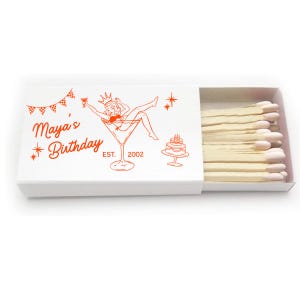 May include: A white matchbox with a red illustration of a woman in a martini glass, a cake, and the text "Maya's Birthday EST. 2002". The matchbox is open, revealing a row of matches with white tips.