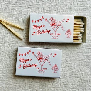 May include: Two white matchboxes with red text and illustrations, one open to reveal matches. The text reads "Maya's Birthday" with a martini glass illustration and a cake. Two matches are next to the boxes.