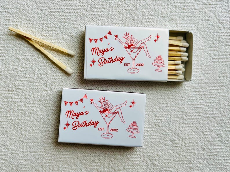 May include: Two white matchboxes with red text and illustrations, one open to reveal matches. The text reads "Maya's Birthday" with a martini glass illustration and a cake. Two matches are next to the boxes.