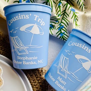 May include: Two light blue plastic cups with white text and graphics. The cups are labeled "Cousins' Trip" and feature a beach scene with a chair, umbrella, and seagulls. One cup says "Stephanie Outer Banks, NC" and the other says "Matt Outer Banks".