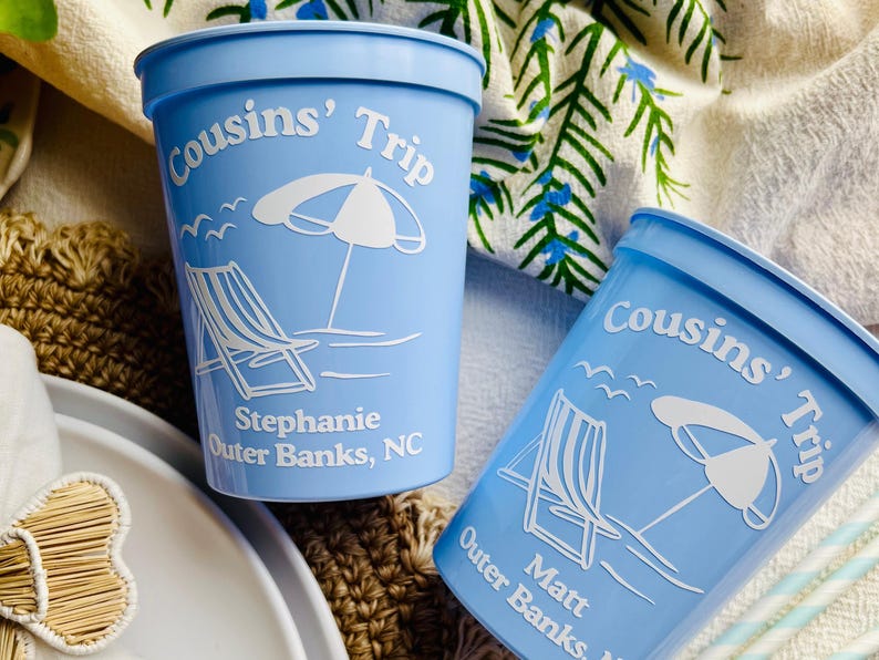 May include: Two light blue plastic cups with white text and graphics. The cups are labeled "Cousins' Trip" and feature a beach scene with a chair, umbrella, and seagulls. One cup says "Stephanie Outer Banks, NC" and the other says "Matt Outer Banks".