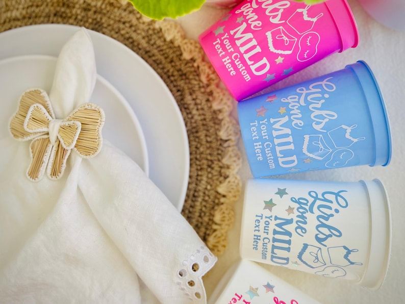 May include: A table setting featuring white plates, a white napkin with a woven bow napkin ring, and three plastic cups. The cups are pink, blue, and white, with the text "Girls gone Mild" and "Your Custom Text Here".
