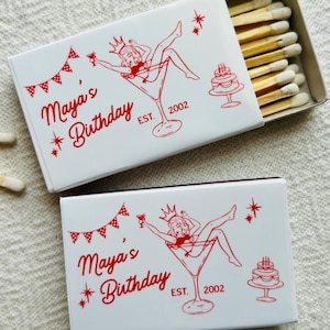 May include: Two white matchbox sets with red graphics. Each box features the text "Maya's Birthday" and a pin-up girl in a martini glass. One box is open, revealing wooden matches. The design includes a banner, cake, and the year "2002".