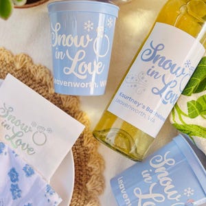 May include: A collection of party supplies with a winter theme. Includes two light blue plastic cups, a wine bottle with a label, and a white napkin. All items feature the text "Snow in Love" in a script font.
