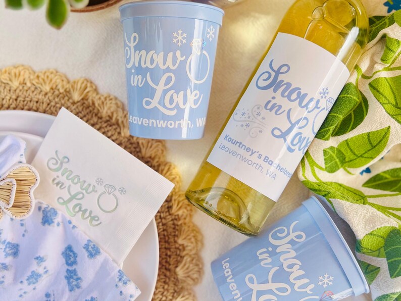 May include: A collection of party supplies with a winter theme. Includes two light blue plastic cups, a wine bottle with a label, and a white napkin. All items feature the text "Snow in Love" in a script font.