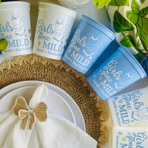 May include: White and light blue plastic cups with the text "Girls gone MILD" and a lingerie graphic. The cups are arranged with white plates, a napkin, and a bow-shaped napkin ring. A decorative placemat is also visible.