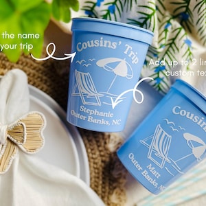May include: Two light blue plastic cups with white text and beach-themed graphics. The cups read "Cousins' Trip" and include a beach chair, umbrella, and custom text. One cup says "Stephanie Outer Banks, NC" and the other says "Matt Outer Banks, NC".