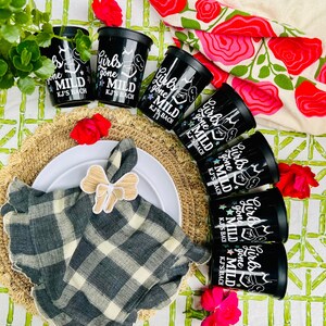 May include: Black cups with the text "Girls gone MILD KJ'S BACH" are arranged around a plate with a napkin and bow. The scene includes a woven placemat, flowers, and a floral tablecloth. The cups are black.