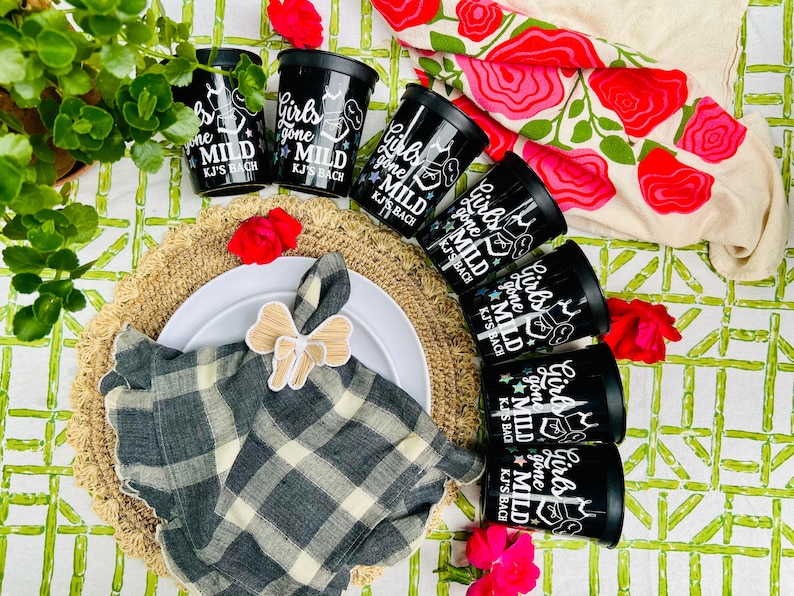 May include: Black cups with the text "Girls gone MILD KJ'S BACH" are arranged around a plate with a napkin and bow. The scene includes a woven placemat, flowers, and a floral tablecloth. The cups are black.