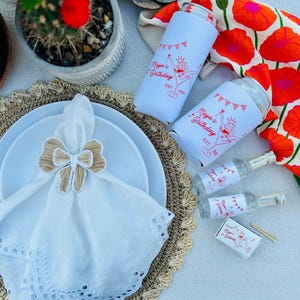 May include: A festive table setting with white plates, a white napkin with a bow-shaped napkin ring, and two white drink koozies with a red graphic that says "Maya's Birthday". Also, there are small bottles and a matchbox with the same graphic.