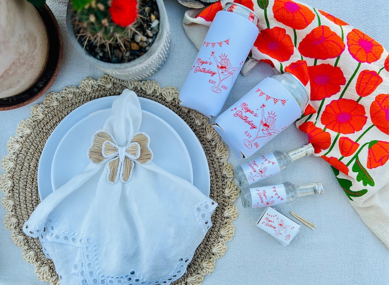 May include: A festive table setting with white plates, a white napkin with a bow-shaped napkin ring, and two white drink koozies with a red graphic that says "Maya's Birthday". Also, there are small bottles and a matchbox with the same graphic.