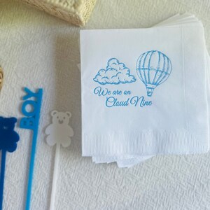 Op de afbeelding: Een stapel witte papieren servetten met een blauw ontwerp van een luchtballon en een wolk. De servetten hebben de tekst "We are on Cloud Nine" erop gedrukt.