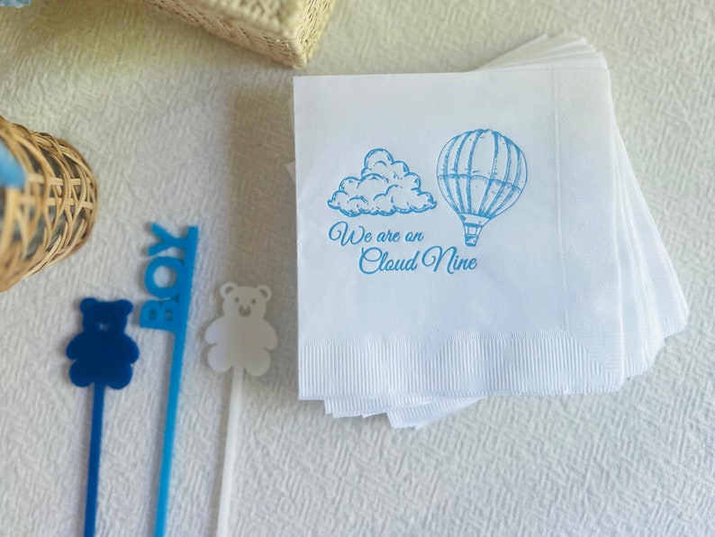 Op de afbeelding: Een stapel witte papieren servetten met een blauw ontwerp van een luchtballon en een wolk. De servetten hebben de tekst "We are on Cloud Nine" erop gedrukt.