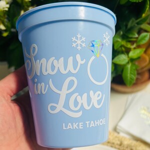 May include: A light blue plastic cup with the words "Snow in Love" in white script, along with a diamond ring graphic and snowflakes. The text "Lake Tahoe" is printed at the bottom.