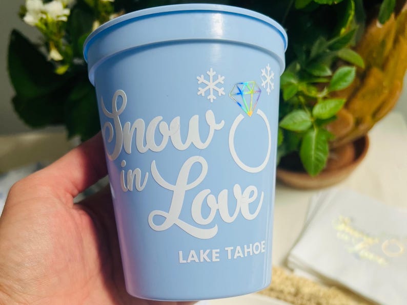 May include: A light blue plastic cup with the words "Snow in Love" in white script, along with a diamond ring graphic and snowflakes. The text "Lake Tahoe" is printed at the bottom.
