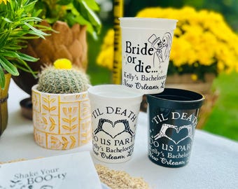 May include: Four plastic cups with text and graphics. Two white cups have the phrases "Bride or die" and "Til Death Do Us Part" with skeleton designs. A black cup also says "Til Death Do Us Part". The cups are for a bachelorette party.