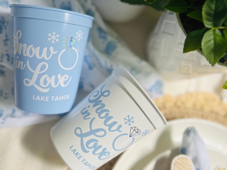 May include: Two plastic cups, one light blue and one white, with the words "Snow in Love Lake Tahoe" printed in a script font. The design includes snowflakes and a diamond ring graphic. The cups are likely for a wedding or event.