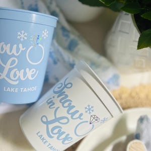 May include: Two plastic cups, one light blue and one white, with the words "Snow in Love" in cursive, a snowflake, and a diamond ring graphic. The text "Lake Tahoe" is printed below. The cups are for a wedding or event.