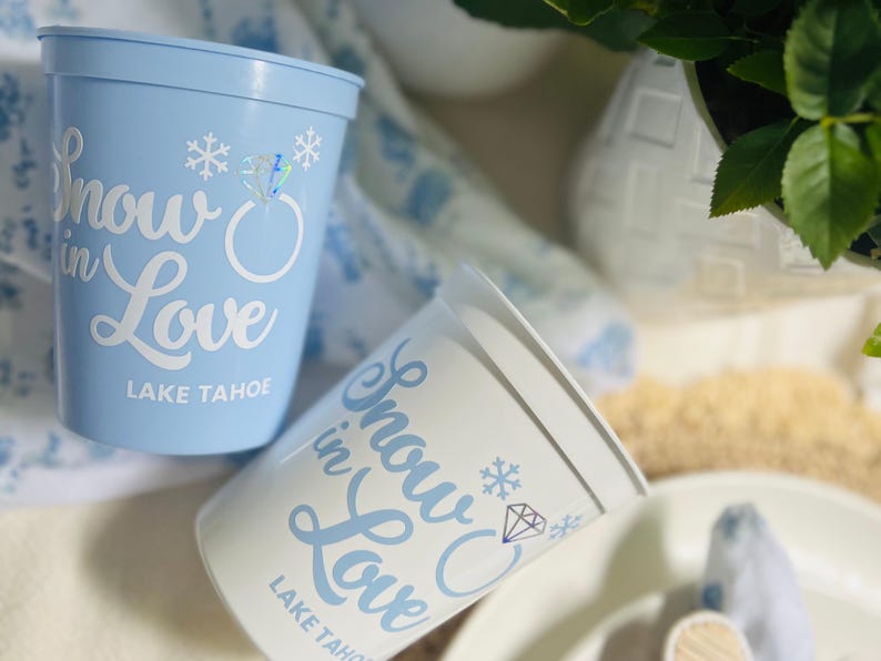 May include: Two plastic cups, one light blue and one white, with the words "Snow in Love" in cursive, a snowflake, and a diamond ring graphic. The text "Lake Tahoe" is printed below. The cups are for a wedding or event.