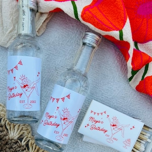 May include: Two clear glass bottles and a matchbox with red and white labels that read "Maya's Birthday EST. 2002". The bottles are next to a floral patterned cloth with red flowers and green stems.