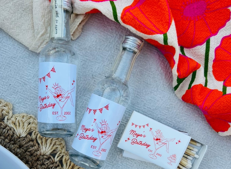 May include: Two clear glass bottles and a matchbox with red and white labels that read "Maya's Birthday EST. 2002". The bottles are next to a floral patterned cloth with red flowers and green stems.