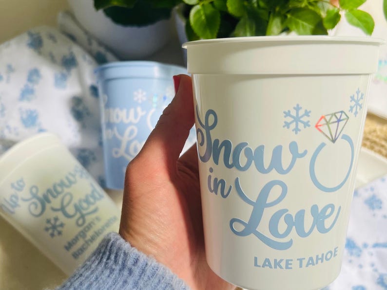 May include: White plastic cup with the words "Snow in Love" in light blue script, along with a diamond ring graphic and snowflakes. The text "Lake Tahoe" is below. Additional cups are visible.
