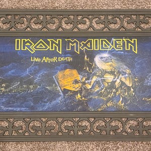 May include: Black doormat with an ornate design featuring a black and white image of a rock band, Iron Maiden, with the text "Live After Death".