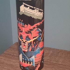 May include: A black stainless steel tumbler with a graphic of a red and blue monster with the text "Defenders of the Faith Judas Priest" on the front.