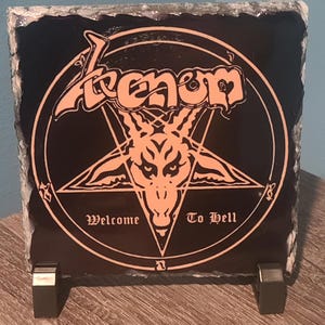 May include: Black square slate featuring the word "Venom" in orange gothic lettering, with a pentagram and a goat's head. The text "Welcome To Hell" is below the pentagram. The slate is displayed on a metal stand.