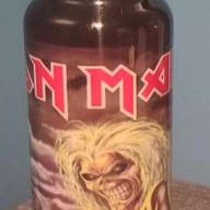 May include: A stainless steel water bottle featuring the band Iron Maiden. The bottle has a dark brown background with the band's logo in red and an illustration of the band's mascot, Eddie. The lid is silver with a wooden top and a silver handle.