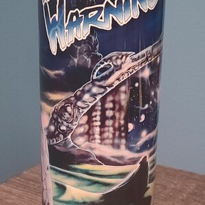 May include: A cylindrical tumbler featuring the words "Fates Warning" in a stylized font against a dark, fantasy-themed backdrop. The design includes a cityscape, a serpent-like creature, and a starry sky.