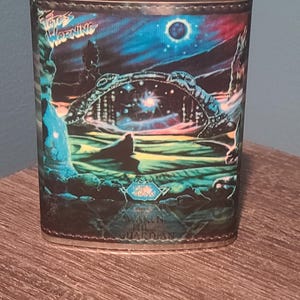 May include: Stainless steel flask with a colorful fantasy art design. The artwork features a celestial scene with the words "Fates Warning" and "Awaken the Guardian". The flask has a silver cap and a leather-like wrap.