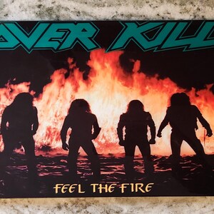 May include: A black and green album cover with the band name "OVERKILL" in large, white letters. The album cover features four silhouettes of people standing in front of a large fire. The album title "FEEL THE FIRE" is written in gold letters at the bottom of the cover.