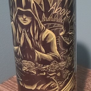 May include: A stainless steel tumbler with a black and gold design. The design features a hooded figure with a crown and the words "Above the rest".