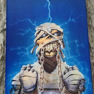 May include: A blue background with a white lightning bolt. A mummy with a chain around its neck and wrists is in the center of the image. The mummy has a white bandage wrapped around its head and face. The mummy's eyes are glowing red.