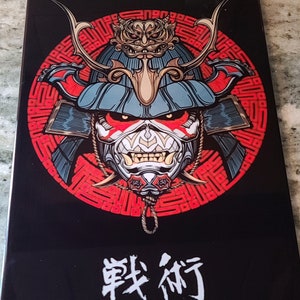May include: A black and red illustration of a samurai warrior wearing a helmet with a fierce expression. The warrior is surrounded by a red and white circular pattern. The text "戦術" is written in white below the warrior.