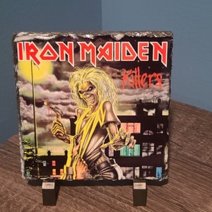 May include: A black slate coaster with a colorful image of the Iron Maiden album cover for "Killers". The image features a yellow-skinned figure with a skull face and a red-stained knife.